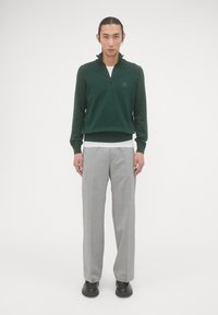 Groene pullover met een ritskraag, gemaakt van zacht materiaal, gecombineerd met lichtgrijze, op maat gemaakte broeken en zwarte schoenen.