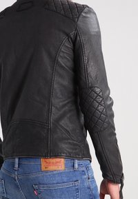 Giacca in pelle nera con dettagli trapuntati su spalle e gomiti, design aderente e cuciture visibili. Indossata sopra jeans blu con marchio.