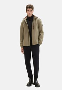 Beige hooded jacket with zipper and red accents, worn over a black turtleneck. Paired with black slim-fit pants and black ankle boots.