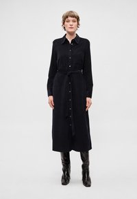 Tmavě modré šaty-košile s dlouhým rukávem, zapínáním na knoflíky vpředu, látkovým páskem a náprsní kapsou. Nošené s černými kozačkami ke kolenům. Jemná textura.