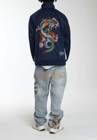 Marineblauwe jas met ingewikkelde draak-epauletten in levendige kleuren; lichtblauwe jeans met bloemen- en schedeldetails; witte sneakers met ontwerpen.