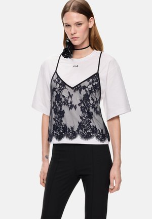 Donna che indossa una maglietta bianca oversize sovrapposta a una canottiera di pizzo nera, pantaloni neri e un choker nero con un dettaglio floreale.