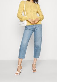 Pull jaune en tricot avec manches bouffantes, associé à un jean court bleu clair et des talons blancs à fines lanières. Un sac bandoulière blanc apporte du contraste.