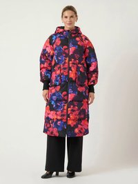 Kvinne iført en lang, hettejakke med puffermønster i lyse røde, rosa og blå blomster, svarte vide bukser og svarte hæler.