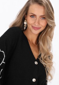 Fekete kardigán puha anyagból, V nyakkal, három ezüst gombbal és fehér virágmintával a bal vállon.