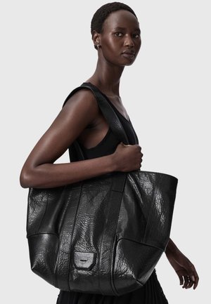 Femme aux cheveux courts portant un haut noir sans manches, avec un grand sac cabas en cuir noir texturé sur l'épaule.