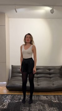 Jeune femme debout, yeux fermés, dans un salon, portant un débardeur beige, un short noir et des collants noirs, devant un canapé gris foncé.