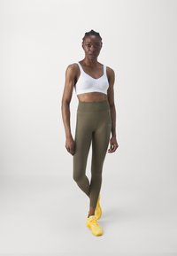 Sports-bh i hvit med teksturert stoff, kombinert med olivengrønne high-waisted leggings. Gule treningssko fullfører antrekket.