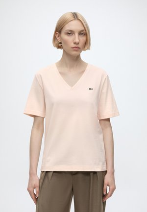 Femme aux cheveux blonds courts portant un T-shirt rose clair à col en V avec un petit logo sur la poitrine et un pantalon marron sur fond uni.