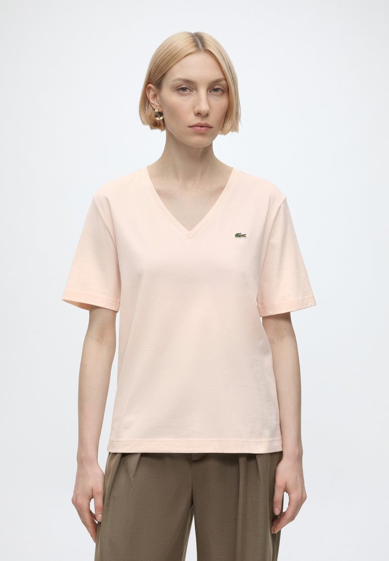 Femme aux cheveux blonds courts portant un T-shirt rose clair à col en V avec un petit logo sur la poitrine et un pantalon marron sur fond uni.