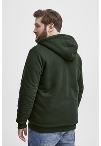 Sudadera verde oscuro con una textura suave y lisa y capucha plana. El diseño cuenta con puños y dobladillo acanalados, combinada con jeans azules.