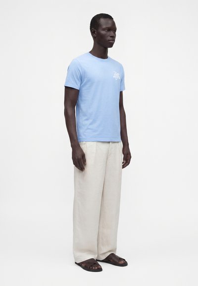 Homme debout portant un t-shirt bleu clair à manches courtes avec un petit motif de tortue blanche, un pantalon ample beige et des sandales marron foncé.