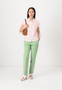 Camisa rosa com riscas e mangas curtas, calças verdes, sandálias brancas de salto bloco, a segurar uma mala castanha. Design simples, ajuste personalizado.