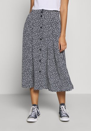 Femme portant une jupe midi fleurie noire et blanche avec des boutons sur le devant, associée à des baskets noires montantes et des chaussettes blanches.