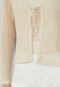 Beige gestrickte Strickjacke mit einem strukturierten, offenen Webdesign. Hat einen Bindeschluss am Ausschnitt und lange Ärmel. Getragen über einem leichten Oberteil.