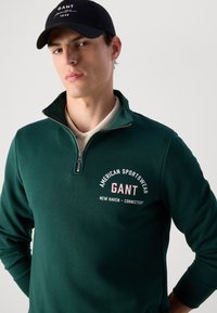 Mężczyzna w ciemnozielonej bluzie typu quarter-zip z napisem "GANT American Sportswear" i czapce baseballowej GANT w kolorze czarnym.