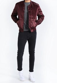 Burgunderfarbene Satinspringjacket mit geripptem Kragen, Fronttaschen, grauem Pullover, schwarzen Jeans und weißen Turnschuhen, vor einem hellen Hintergrund.
