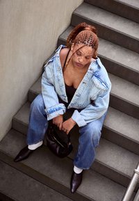 Mlada ženska s pletenicami in pegicami sedi na stopnicah, oblečena v jeans jakno in hlače, v rokah drži črno torbico.