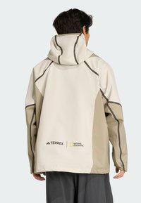Letvægts beige og hvid jakke med hætte, udstyret med kontrastfarvede grå sømme og logoer for Terrex og National Geographic.