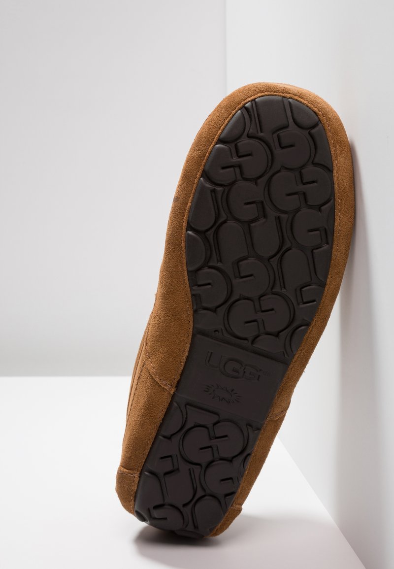 Brown suede slipper with a rubber sole featuring an embossed UGG logo pattern, angled against a white background.