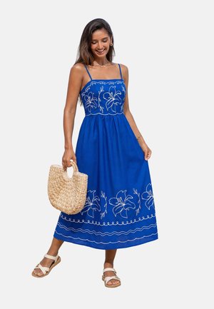 Mujer con un vestido azul sin mangas con bordados florales blancos, sosteniendo un bolso tejido y con sandalias blancas.