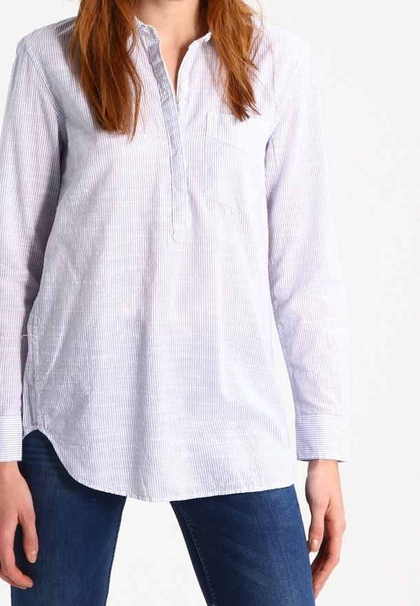 Personne portant une chemise à manches longues à rayures bleu clair avec une patte de boutonnage partielle à l’avant et un jean bleu foncé, debout devant un fond blanc.