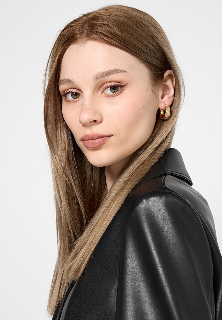 Schwarze Lederjacke mit glänzender Oberfläche. Model mit langen, geraden, hellbraunen Haaren, das auffällige, strukturierte goldene Creolen trägt.