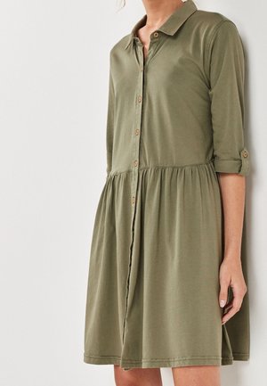 Kvinde iført olivengrøn knap-op-kjole med indsnævret talje og op rullede ærmer, stående mod en ensformig hvid baggrund.