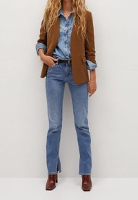 Blazer marron porté sur une chemise en denim clair, associé à un jean bleu avec des fentes latérales. Des bottines marron �à talons complètent la tenue.