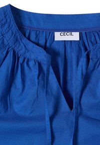 Blouse en coton bleu avec encolure froncée et ouverture devant en forme de V, comportant une étiquette blanche avec le texte "CECIL" à l'intérieur du col.