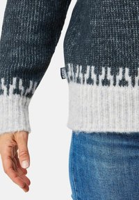 Sweter dzianinowy w ciemnoszarym kolorze z białym wzorzystym wykończeniem u dołu i przy mankietach. Posiada miękki, teksturowany materiał oraz widoczną metkę z logo po boku.