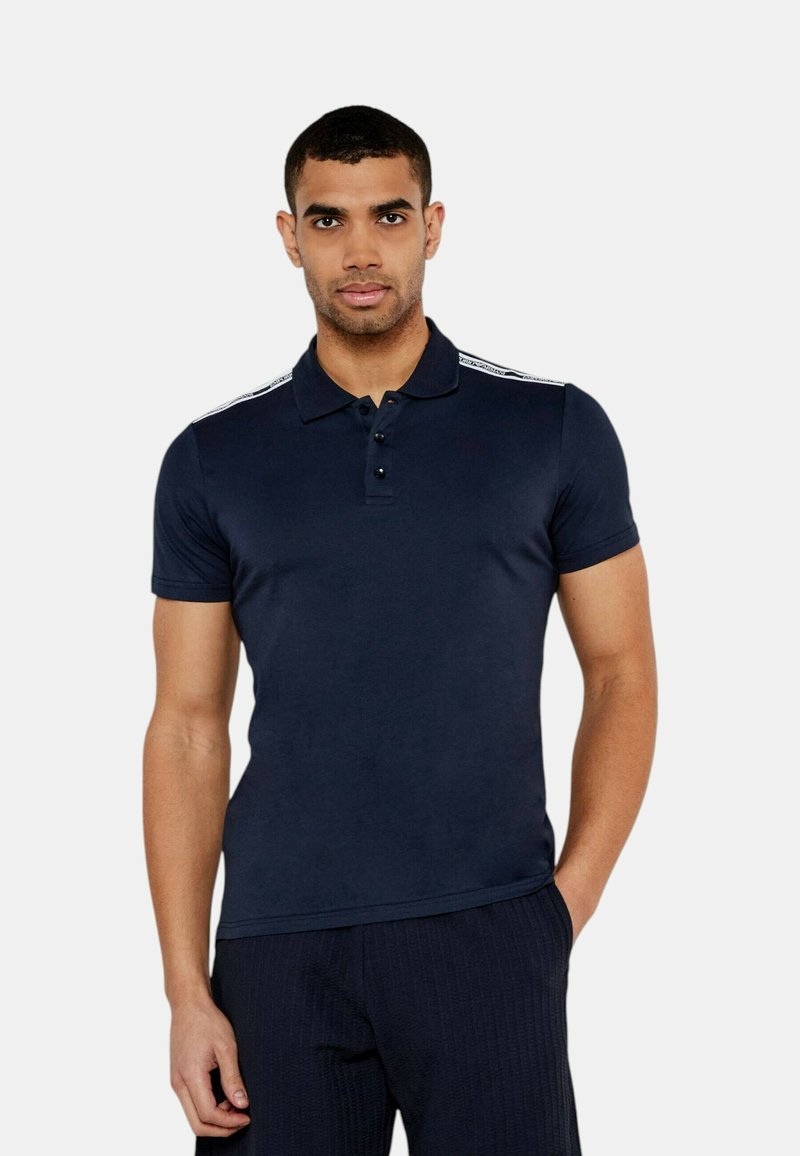 Polo shirt blu navy in un tessuto liscio con maniche corte, patta con tre bottoni e strisce bianche a contrasto lungo le spalle.