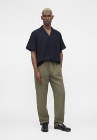 Homme aux cheveux blonds courts portant une chemise noire à manches courtes boutonnée, un pantalon vert olive et des chaussures habillées noires, debout sur fond blanc.