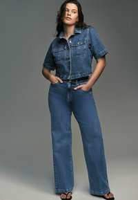 Veste en denim à manches courtes et deux poches avant, associée à un jean taille haute à jambes larges, le tout dans un bleu moyen. Sandales noires.