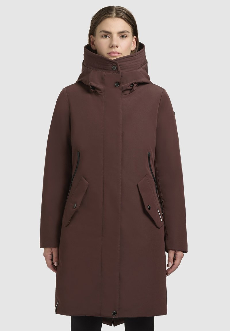 Cappotto marrone fino al ginocchio con colletto alto, zip frontale e grandi tasche laterali. Tessuto liscio, opaco con una leggera trama e dettagli in contrasto.