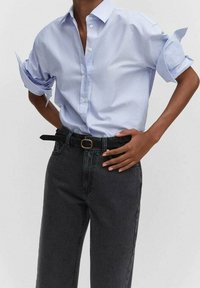 Camisa azul claro de botones con las mangas remangadas, combinada con jeans de mezclilla oscura de talle alto y un cinturón negro con una hebilla metálica redonda.