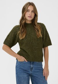 Moteris su ilgais banguotais plaukais, aps wearing olive green trumpas rankoves megztuką ir mėlynas džinsus, pozuojanti su viena ranka kišenėje.