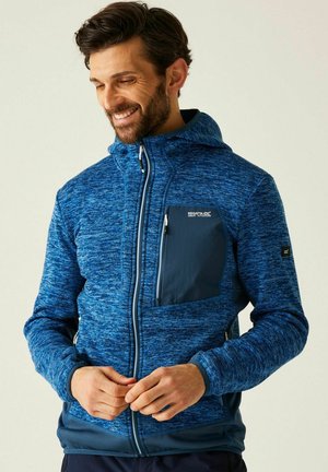 Mężczyzna uśmiechający się podczas zapinania niebieskiej kurtki z kapturem z teksturowanego materiału z kieszenią na piersi na gładkim jasnym tle.