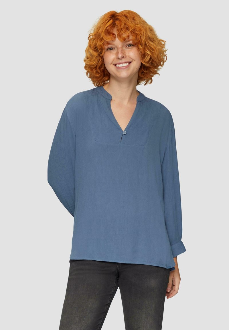 Lichtblauwe blouse met lange mouwen en een notch-kraag, voorzien van een borstzak en subtiele textuur. Gecombineerd met donkergrijze jeans.