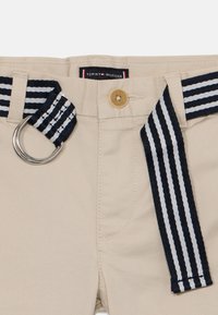Pantalon beige avec une finition texturée, équipé d'une ceinture à rayures bleu marine et blanc, d'un anneau en D en métal et d'une fermeture par bouton portant l'inscription "Tommy Hilfiger".