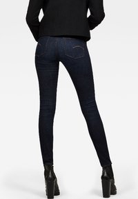 Jeans ajustados de mezclilla oscura con una textura suave, corte slim, bolsillos traseros y detalles de costura sutiles, combinados con botas de tacón negras.