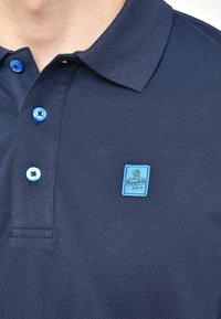 Námořnické polo tričko s texturovanou látkou, které má světle modré knoflíky a modrou nášivku na levé části hrudi.