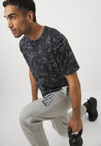 Sort camouflage-mønstret T-shirt med korte ærmer, grå joggebukser med logo-detalje, sort kettlebell og atletiske sko i en træningsstilling.