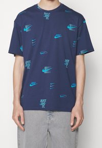 Navy blue cotton t-shirt featuring turquoise Nike logos and "Just Do It" text in a scattered pattern. Short sleeves and round neckline.