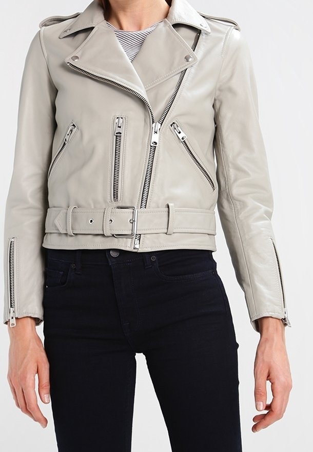 Veste motard en cuir gris clair avec de larges revers, plusieurs fermetures éclair argentées et une ceinture assortie portée avec un pantalon noir.