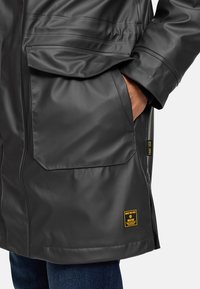 Abrigo negro impermeable con dos grandes bolsillos frontales, textura suave, costuras visibles y etiqueta de marca amarilla en la esquina inferior derecha.
