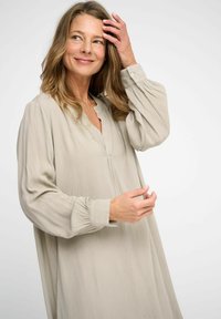 Long-sleeve, light beige dress with a relaxed fit, V-neckline, and softly gathered cuffs; made from a textured fabric, smooth appearance.