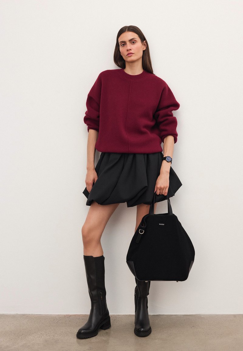 Burgundi pulóvert, fekete puffos szoknyát, térd fölé érő fekete csizmát viselő nő, aki egy nagy fekete táskát tart, és egy egyszerű világos fal előtt áll.