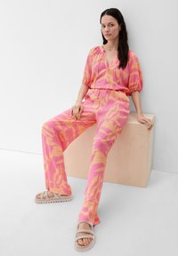 Roze en oranje gepatineerde tweedelige outfit met een top met ballonmouwen en een wijde broek, aangevuld met lichtgewicht, bandjesbeige sandalen.