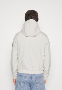 Light gray hooded jacket with a smooth texture, elastic cuffs, and waistband. Features a logo patch on the left shoulder. Denim jeans visible below.